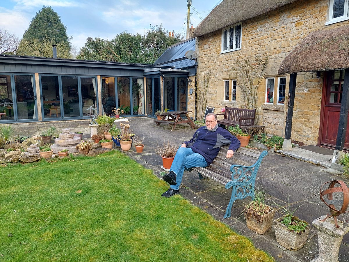 Tim sat on a bench in the garden of his grade II listed home with a thatched roof. Solar panels are on the extension of the home.