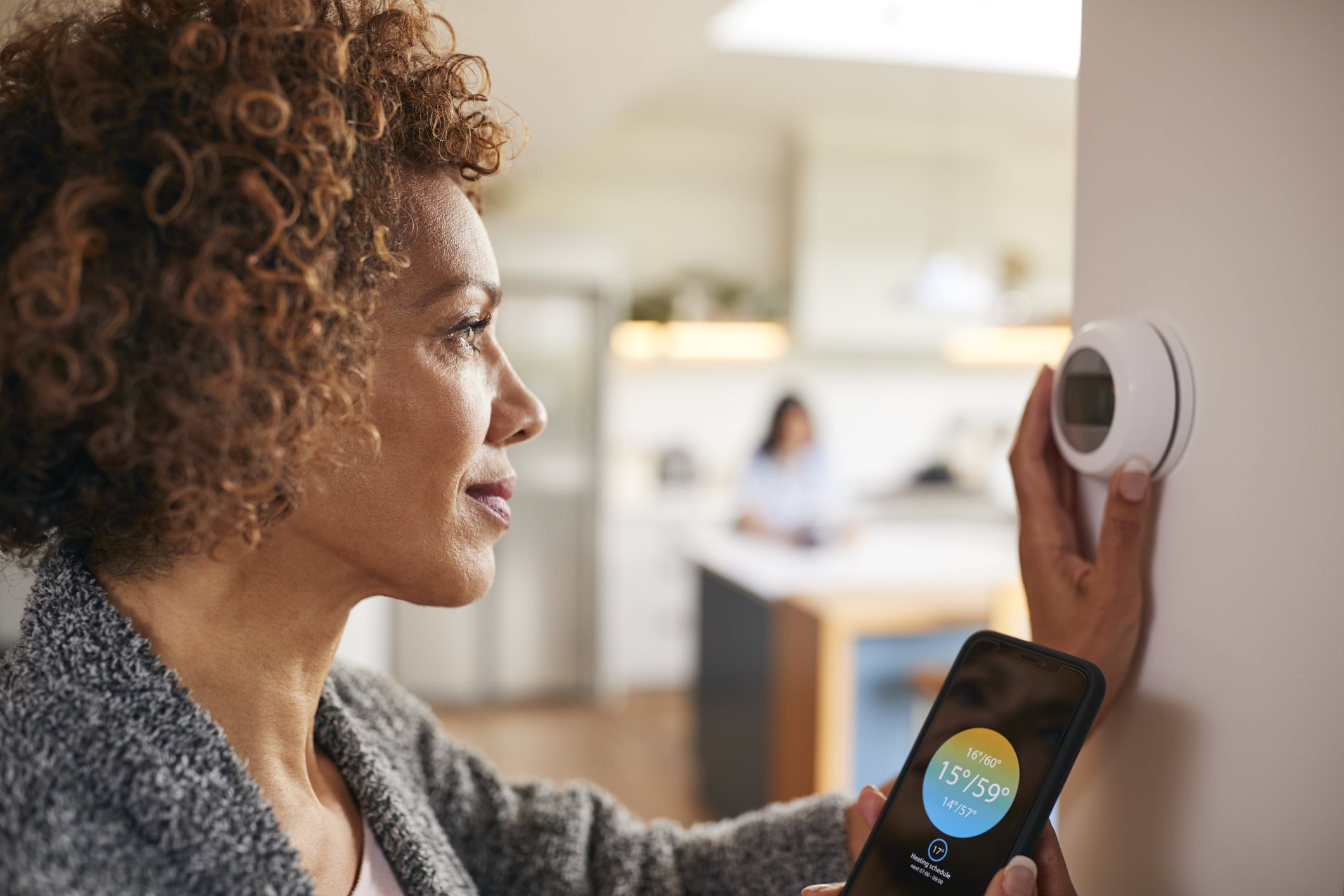 A woman adjusting a digital central heating system thermostat while holding a smartphone showing the corresponding smart heating control app.