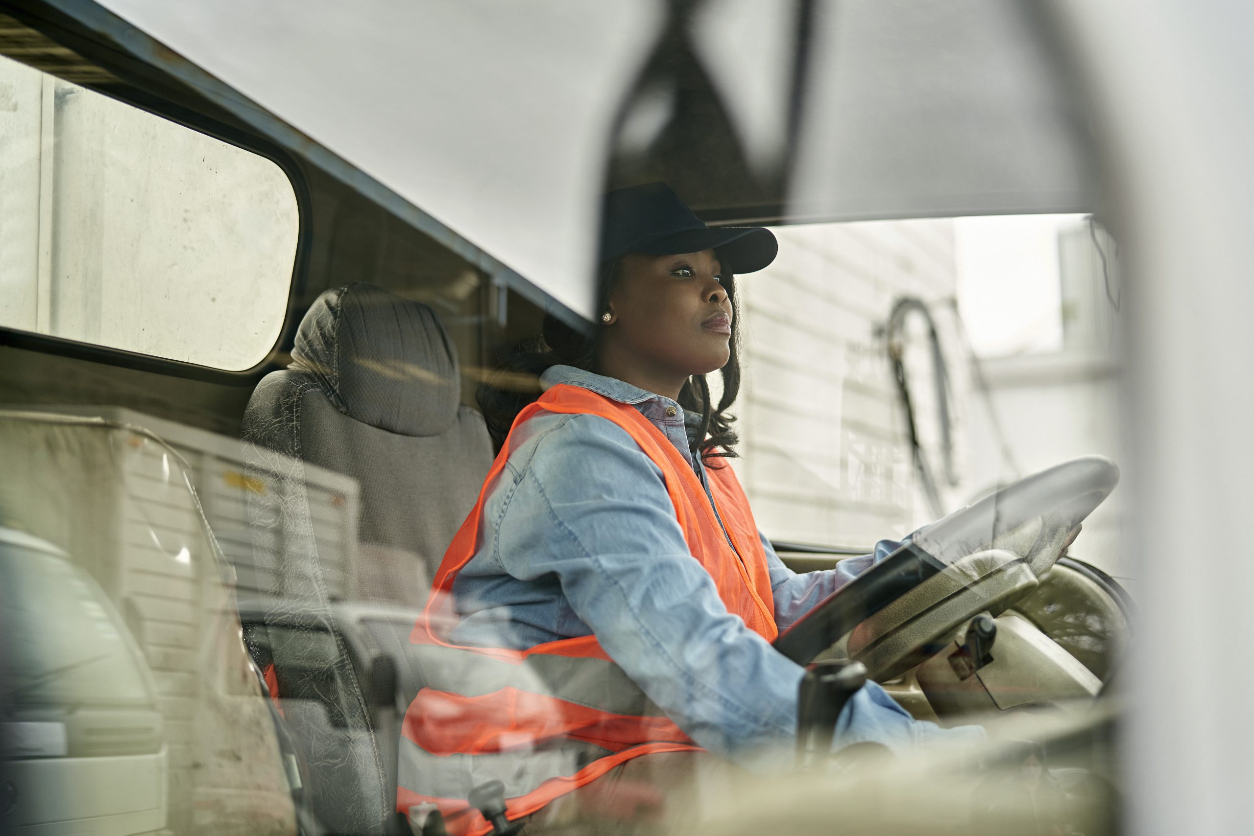 Personal perspective of mid 20s woman in casual clothing, cap, and reflective vest sitting in driver’s seat HGV