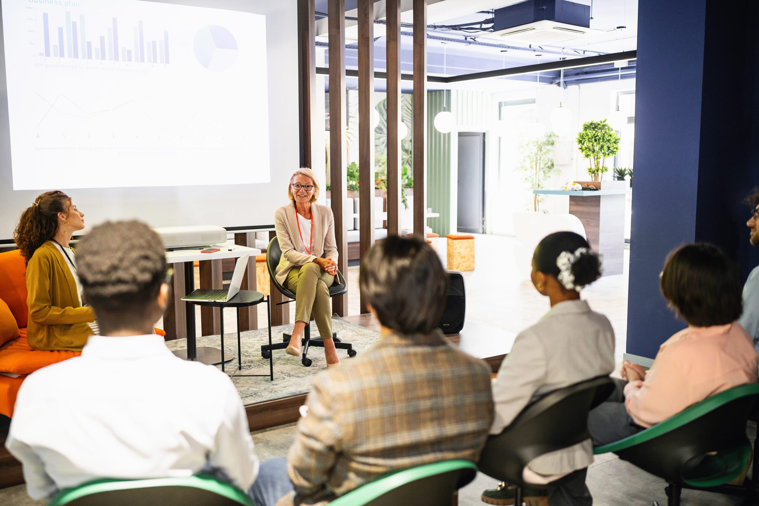 Two women sitting on a stage presenting to a group of colleagues sitting on chairs in the audience at an office